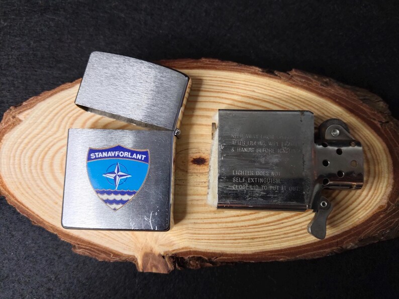 Może przedstawiać: Srebrna zapalniczka z otwartą pokrywą, prezentująca niebiesko-biały emblemat z napisem "STANAVFORLANT". Zapalniczka leży na drewnianej powierzchni. Tekst wewnątrz brzmi "KEEP AWAY FROM CHILDREN" i "LIGHTER DOES NOT SELF EXTINGUISH".