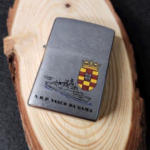 Może przedstawiać: Srebrna zapalniczka ze szczotkowanym metalowym wykończeniem. Zapalniczka posiada szczeg&oacute;łową ilustrację statku, helikoptera i herbu. Tekst "N.R.P. VASCO DA GAMA" jest wydrukowany pod ilustracją statku.