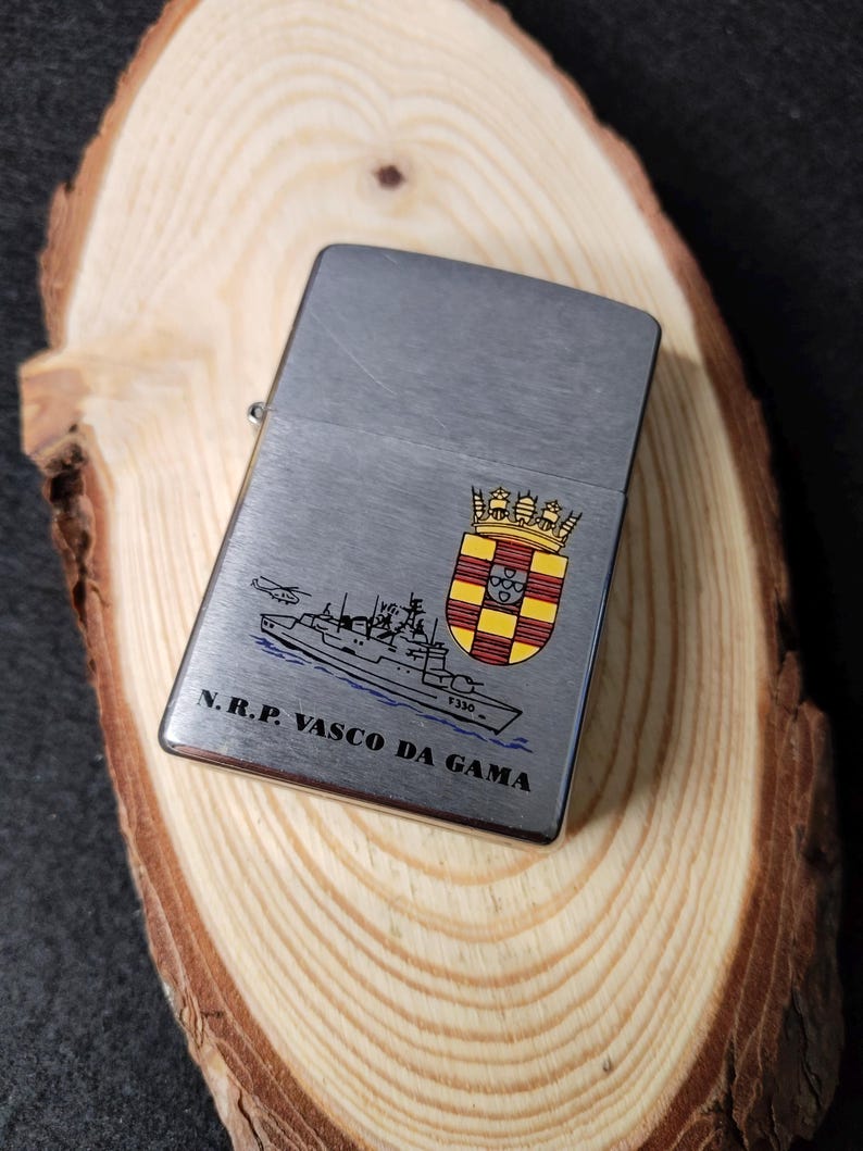 Może przedstawiać: Srebrna zapalniczka ze szczotkowanym metalowym wykończeniem. Zapalniczka posiada szczeg&oacute;łową ilustrację statku, helikoptera i herbu. Tekst "N.R.P. VASCO DA GAMA" jest wydrukowany pod ilustracją statku.