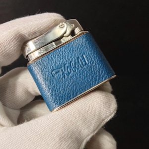 Puede incluir: Un encendedor vintage con exterior de cuero azul y detalles metálicos plateados. El encendedor tiene la palabra "Vokold" grabada en el cuero azul. El encendedor es rectangular con una tapa con bisagras.
