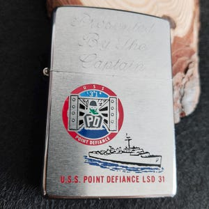 Puede incluir: Un encendedor de metal plateado con la inscripción "Presented By The Captain". Presenta un emblema colorido con "USS 31" y "PD", y un dibujo de un barco. También se puede ver el texto "U.S.S. POINT DEFIANCE LSD 31".