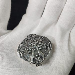 Puede incluir: Un broche de metal plateado con un diseño en relieve de un rostro central rodeado de cabello fluido y motivos florales. La pieza tiene una forma redondeada irregular con un acabado pulido que resalta los intrincados detalles esculpidos.