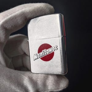 Peut inclure: Un briquet argent&eacute; avec le mot "Manitowoc" en blanc sur un cercle rouge. Le briquet est tenu dans une main gant&eacute;e de blanc sur un fond sombre.