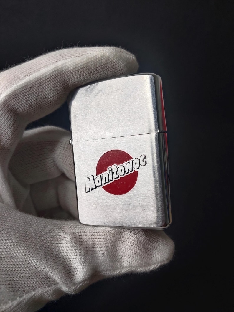 Peut inclure: Un briquet argent&eacute; avec le mot "Manitowoc" en blanc sur un cercle rouge. Le briquet est tenu dans une main gant&eacute;e de blanc sur un fond sombre.