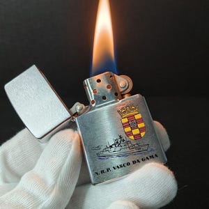 Może przedstawiać: Srebrna metalowa zapalniczka z płonącym płomieniem. Zapalniczka posiada ilustrację statku i napis "N.R.P. VASCO DA GAMA" wraz z herbem. Zapalniczka jest otwarta, ukazując płomień.