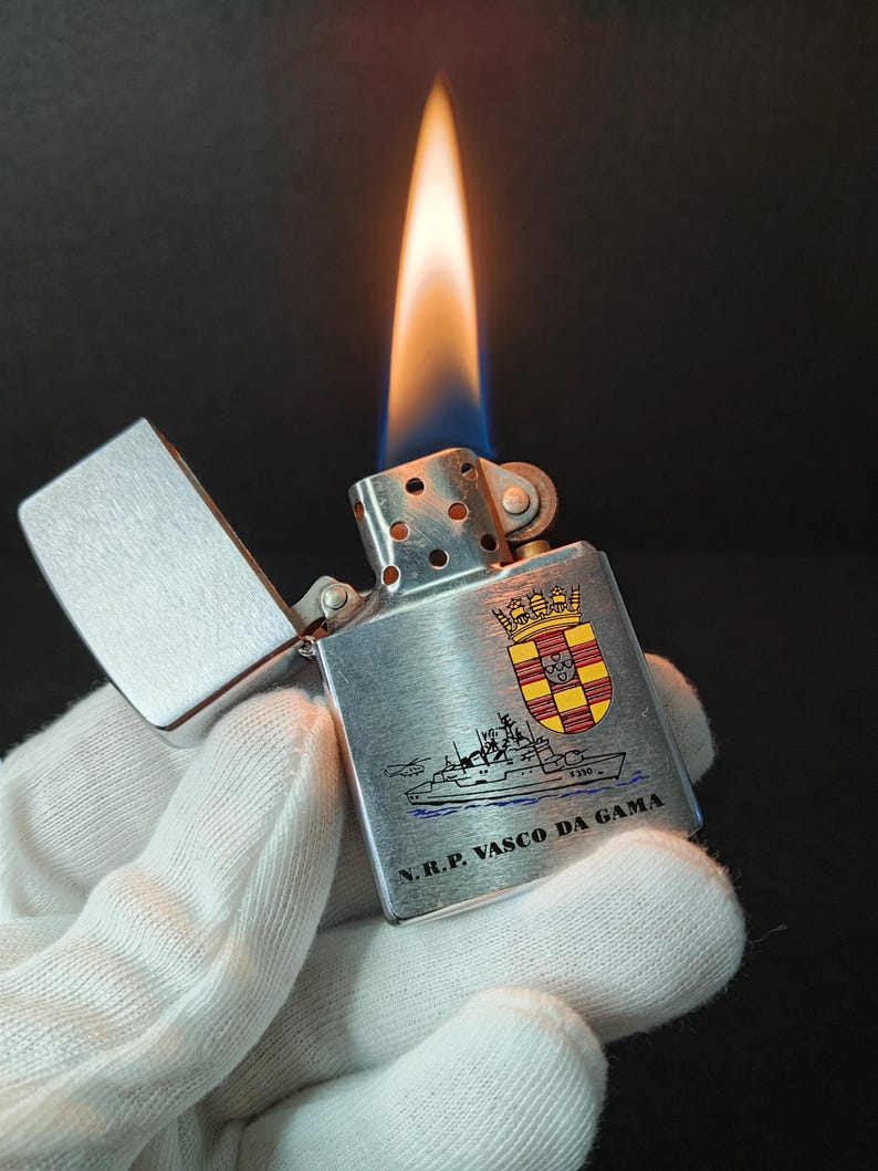 Może przedstawiać: Srebrna metalowa zapalniczka z płonącym płomieniem. Zapalniczka posiada ilustrację statku i napis "N.R.P. VASCO DA GAMA" wraz z herbem. Zapalniczka jest otwarta, ukazując płomień.
