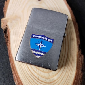 Może przedstawiać: Srebrny metalowy zapalniczka z niebiesko-białą odznaką z napisem "STANAVFORLANT". Na odznace znajduje się gwiazda i faliste linie. Prostokątna zapalniczka leży na drewnianej powierzchni.