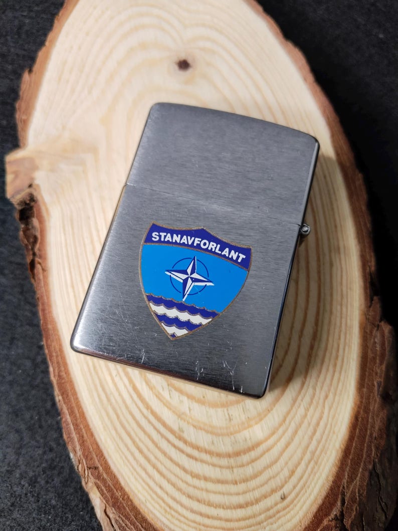 Może przedstawiać: Srebrny metalowy zapalniczka z niebiesko-białą odznaką z napisem "STANAVFORLANT". Na odznace znajduje się gwiazda i faliste linie. Prostokątna zapalniczka leży na drewnianej powierzchni.