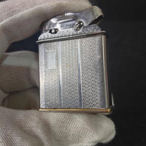May include: A rectangular silver-tone metal lighter featuring a textured, wavy pattern on its surface. The lighter has a smooth, vertical rectangular panel on the left side and a polished metallic top mechanism with visible screws and a hinged lid.
