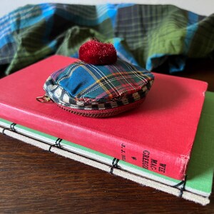 May include: A small, plaid coin purse with a red pom-pom sits on a red book. The purse has a zip and a blue, green, and yellow plaid pattern. A green and blue plaid scarf is in the background.
