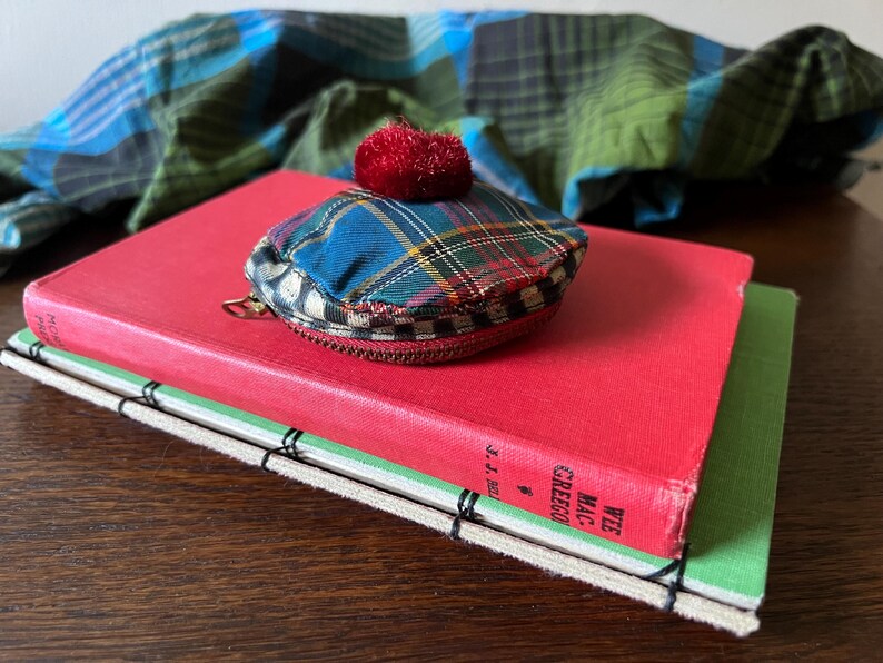 May include: A small, plaid coin purse with a red pom-pom sits on a red book. The purse has a zip and a blue, green, and yellow plaid pattern. A green and blue plaid scarf is in the background.