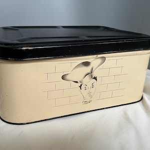 May include: A vintage metal bread box with a black lid and a cream-colored body. The front of the box features a black and white illustration of a chef with a brick wall background.