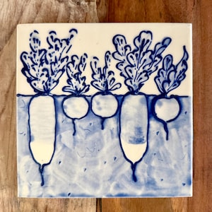 Puede incluir: Azulejo de cerámica cuadrado con un diseño pintado a mano de rábanos en azul y blanco. Los rábanos se representan con sus raíces y hojas, sobre un fondo azul claro.