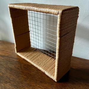 May include: A rectangular, open-front shelf with a woven, light brown rope frame and a silver wire mesh backing. The shelf is designed to be mounted on a wall or placed on a surface. The shelf is made of natural materials.