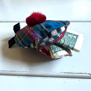 May include: A novelty shark-shaped purse crafted from tartan fabric in blue, green, and red hues. It features a red trim, a red pom-pom, and a black fin. The open mouth of the shark reveals folded dollar bills.