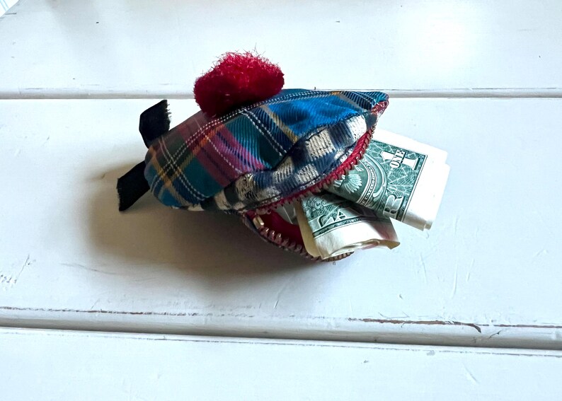 May include: A novelty shark-shaped purse crafted from tartan fabric in blue, green, and red hues. It features a red trim, a red pom-pom, and a black fin. The open mouth of the shark reveals folded dollar bills.