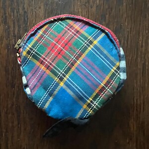 May include: A round coin purse with a tartan pattern in blue, green, red, and yellow. The purse has a black strap and a red trim. The zipper is gold-coloured.