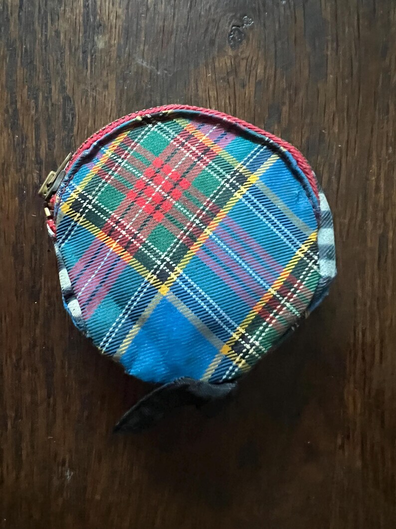May include: A round coin purse with a tartan pattern in blue, green, red, and yellow. The purse has a black strap and a red trim. The zipper is gold-coloured.
