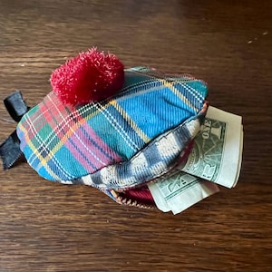 May include: A small, tartan coin purse with a red pom-pom and black ribbon detail. The purse is open, revealing several one-dollar bills. The purse is resting on a wooden surface.