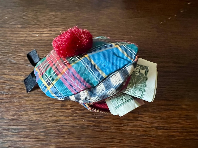 May include: A small, tartan coin purse with a red pom-pom and black ribbon detail. The purse is open, revealing several one-dollar bills. The purse is resting on a wooden surface.