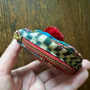 May include: A small, colourful zippered pouch with a red trim and a gold zipper. The pouch features a patterned fabric with blue, green, and brown hues, and a small red pom-pom. The pouch is held in a hand.