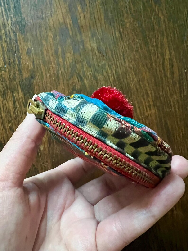 May include: A small, colourful zippered pouch with a red trim and a gold zipper. The pouch features a patterned fabric with blue, green, and brown hues, and a small red pom-pom. The pouch is held in a hand.