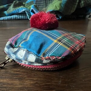 May include: A small, plaid fabric coin purse with a red pom-pom on top. The purse has a zipper closure and a metal clasp. The plaid pattern includes blue, green, red, and yellow. The purse is on a wooden surface.