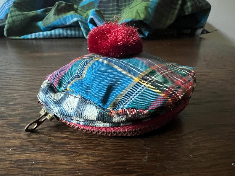 May include: A small, plaid fabric coin purse with a red pom-pom on top. The purse has a zipper closure and a metal clasp. The plaid pattern includes blue, green, red, and yellow. The purse is on a wooden surface.