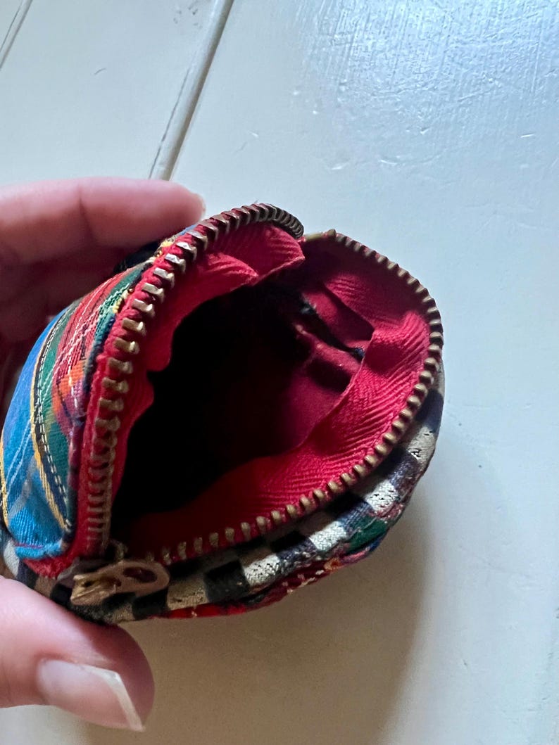 May include: Small, open coin purse with a tartan pattern in red, blue, green, and yellow. The interior is red, and the zipper is gold-coloured. The purse is round, with a vintage aesthetic.