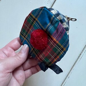 May include: A small, tartan fabric coin purse with a red, textured circle on the front. The purse has a zip closure and a black ribbon detail. The tartan pattern includes blue, green, red, and yellow lines.