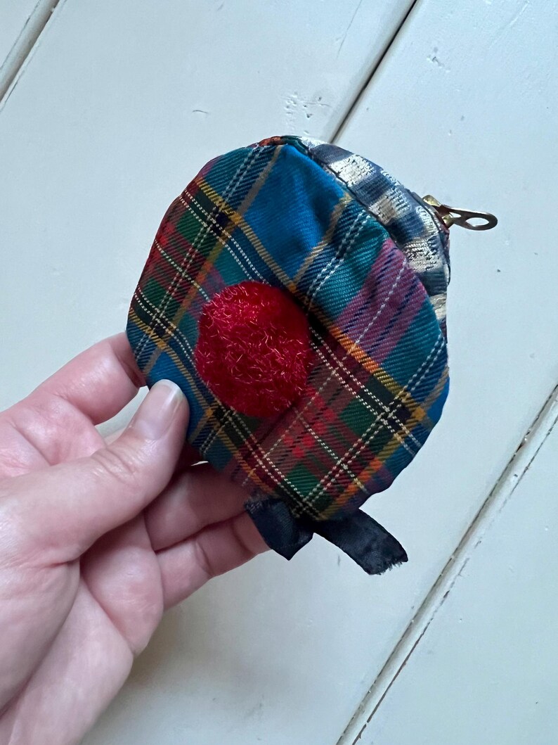 May include: A small, tartan fabric coin purse with a red, textured circle on the front. The purse has a zip closure and a black ribbon detail. The tartan pattern includes blue, green, red, and yellow lines.