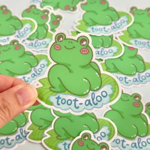May include: Green frog sticker with the text "toot-aloo" in a light blue cloud. The frog is sitting on a green lily pad. The sticker has a white border and is surrounded by other similar stickers.