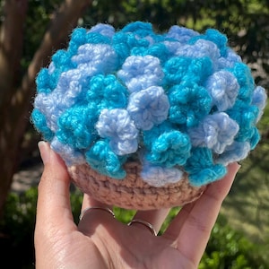 May include: A handmade crochet flower arrangement in a small basket. The flowers are a mix of light blue and turquoise, with a beige basket base. The arrangement is held in a hand, with a blurred green background.