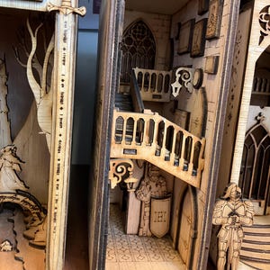 May include: Wooden book nook depicting a medieval castle scene with a staircase, arched doorway, and miniature knight figures. The book nook is crafted from light-coloured wood with intricate laser-cut details.