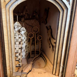 Könnte beinhalten: Ein mehrschichtiges Holzkunstwerk mit einer detaillierten Innenszene. Das Kunstwerk zeigt ein Schloss, einen Besen und Quidditch-Bälle. Das Stück ist aus hellem Holz gefertigt, mit aufwendigen Ausschnitten, die Tiefe und Dimension erzeugen.