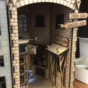 Könnte beinhalten: Ein lasergeschnittenes Holzdiorama, das eine Gasse mit Ziegelmuster und Schildern für Diagon Alley und Knockturn Alley darstellt. Die Struktur umfasst ein Geschäft mit der Aufschrift Borgin and Burkes, ein Vordach, Fenster und einen gepflasterten Boden.
