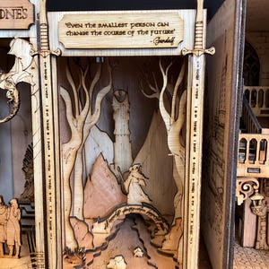 Könnte beinhalten: Ein lasergeschnittenes Holzdiorama mit einer geschichteten Landschaft, einem zentralen Turm, einer Figur mit Stab und einer Brücke. Der Rahmen wird von zwei schwertförmigen Säulen flankiert. Der Text lautet: 'Even the smallest person can change the course of the future' - Gandalf.