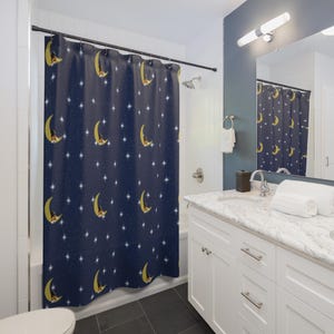 May include: A navy blue shower curtain featuring a repeating pattern of crescent moons with sleeping figures and white stars. The curtain is hanging in a bathroom with white cabinets, a marble countertop, and a mirror.