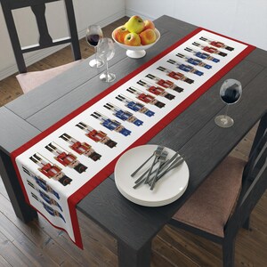 May include: A white table runner with a red border features a repeating pattern of toy nutcracker soldiers in red and blue uniforms. The runner is displayed on a dark gray wooden table, set with plates, silverware, and wine glasses.