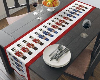 May include: A white table runner with a red border features a repeating pattern of toy nutcracker soldiers in red and blue uniforms. The runner is displayed on a dark gray wooden table, set with plates, silverware, and wine glasses.