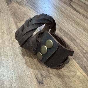 May include: Two brown leather bracelets, one with a braided design and the other with three brass studs.