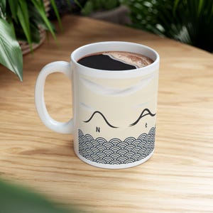Puede incluir: Taza de café de cerámica blanca con líquido negro en su interior. La taza presenta un fondo beige con un diseño de olas negras en la parte inferior y un diseño de líneas negras con las letras "N" y "t".