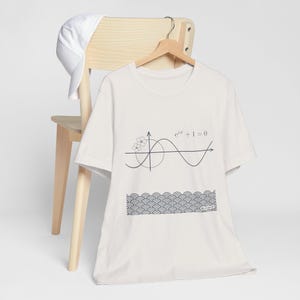 May include: A white t-shirt with a black graphic design featuring a sine wave, a floral element, and the equation e^(iπ) + 1 = 0. Below the equation is a pattern of repeating curved lines. A white baseball cap rests on the back of a wooden chair.