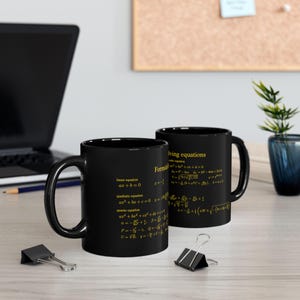 Könnte beinhalten: Zwei schwarze Kaffeetassen mit goldenem Text, der mathematische Formeln anzeigt. Eine Tasse zeigt Gleichungen wie ax+b=0, während die andere Formeln zur Lösung von Gleichungen enthält.