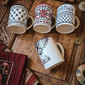 May include: Four white ceramic mugs featuring distinct patterns. Three mugs stand upright, displaying black and white keffiyeh-style prints, a red and black geometric cross-stitch design, and Arabic calligraphy. One mug lies on its side with Arabic text.