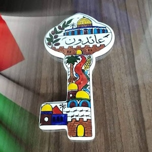 May include: A key-shaped ceramic ornament featuring hand-painted illustrations of the Dome of the Rock, stone walls, olive branches, and palm trees. The Arabic text "عائدون" is inscribed in the center. The design includes yellow domes.