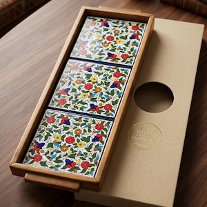 May include: A rectangular wooden serving tray containing three square ceramic tiles with a colorful floral pattern of red, yellow, and blue flowers on a white background. Next to the tray is a tan cardboard box with a gold circular sticker.