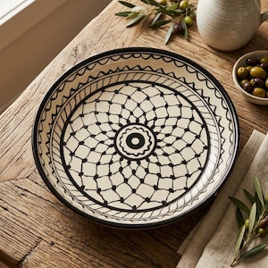 May include: A round ceramic serving bowl featuring a cream-colored base with a hand-painted black geometric pattern. The design consists of a central circular motif surrounded by a repeating net-like lattice and a scalloped border along the rim.