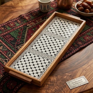 May include: A rectangular wooden serving tray featuring three white ceramic tiles with a black geometric keffiyeh pattern. The tray has integrated handles on the short ends. A small silver-toned metal plaque reads Handcrafted in Palestine.