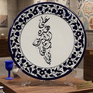 May include: A round ceramic plate featuring a white center with black Arabic calligraphy shaped like a map, surrounded by a dark blue and white floral border. The plate rests on a clear stand, with small labels reading Palestine: A Land of Life.